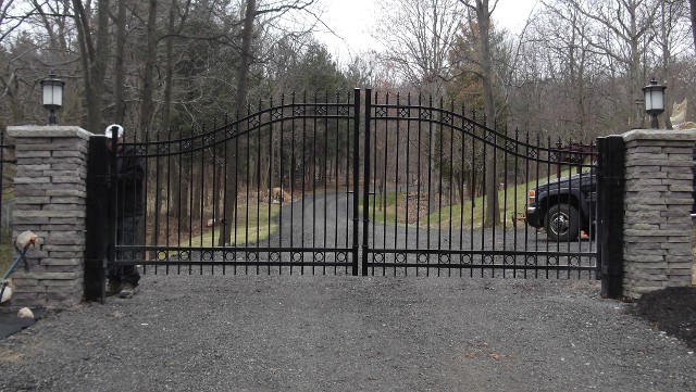 Arched black iron double driveway gate hung between stacked stone pillars with lanterns